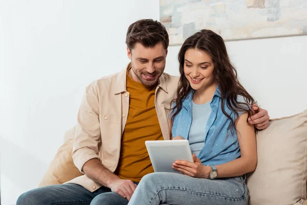 Schöner Mann umarmt lächelnde Freundin, während er zu Hause digitales Tablet benutzt — Stockfoto