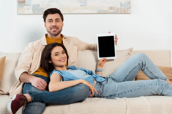 Pareja joven sonriendo a la cámara mientras muestra la tableta digital con pantalla en blanco en la sala de estar - foto de stock