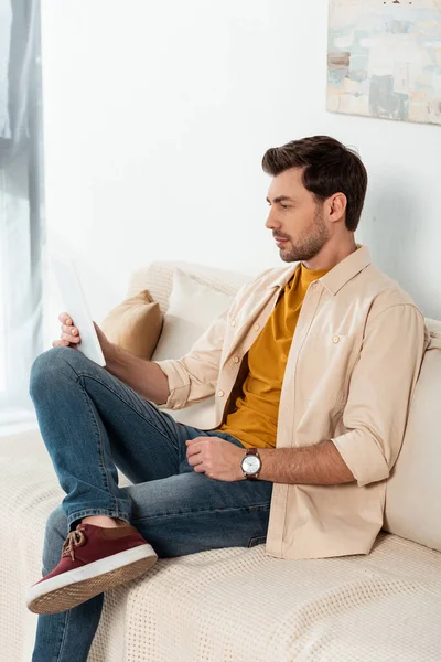Hombre guapo sosteniendo tableta digital en el sofá en la sala de estar - foto de stock