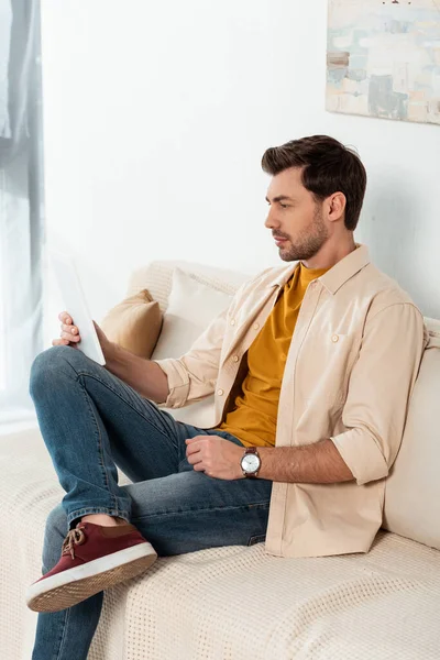 Bonito homem segurando tablet digital no sofá na sala de estar — Fotografia de Stock