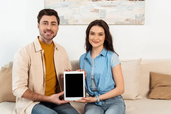 Улыбающаяся пара смотрит в камеру и держит цифровой планшет с пустым экраном на диване — стоковое фото