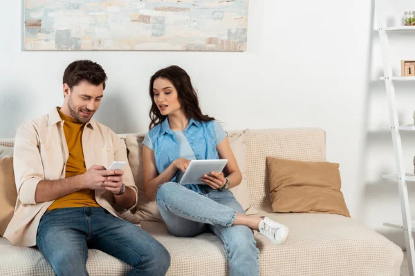 Sorrindo homem usando smartphone perto da namorada com tablet digital em casa — Fotografia de Stock