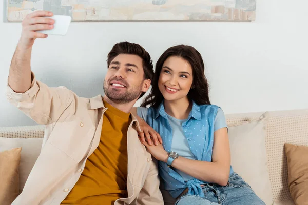 Enfoque selectivo de la pareja sonriente tomando selfie en el teléfono inteligente en la sala de estar - foto de stock