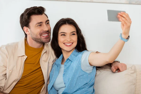 Selective focus of woman taking selfie on smartphone near smiling boyfriend at home — Stock Photo