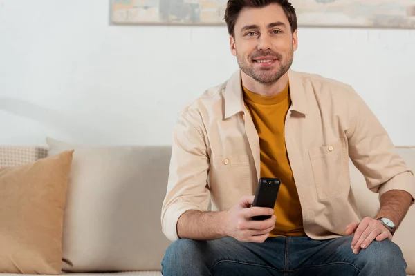 Uomo sorridente che tiene il telecomando mentre guarda la tv a casa — Foto stock