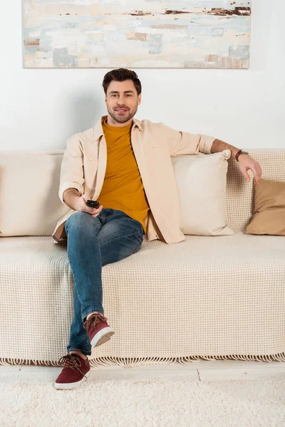 Schöner Mann mit Fernbedienung und lächelnd in die Kamera auf dem Sofa — Stockfoto