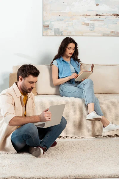 Hombre usando el ordenador portátil cerca de la mujer leyendo libro en la sala de estar - foto de stock