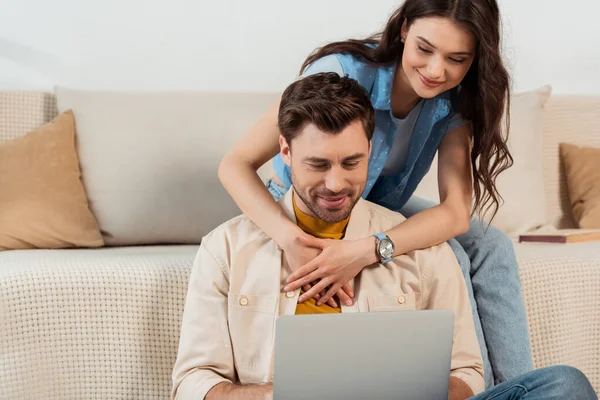 Sonriente chica abrazando novio usando el ordenador portátil en casa - foto de stock