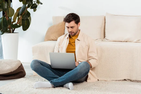 Bonito homem usando laptop no chão perto do sofá — Fotografia de Stock