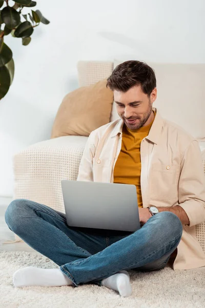 Freelancer sonriente usando portátil mientras está sentado en el suelo en la sala de estar - foto de stock