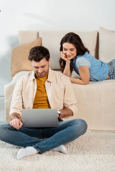Smiling woman lying on couch near boyfriend using laptop on floor — Stock Photo