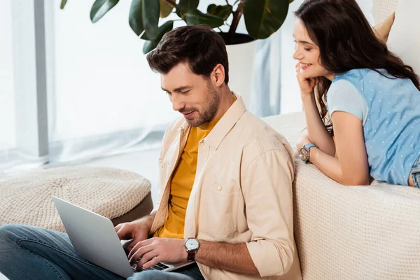 Красивый мужчина, использующий ноутбук рядом с улыбающейся девушкой на диване — стоковое фото