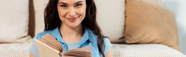 Foto panoramica di una donna sorridente che guarda la macchina fotografica mentre legge un libro a casa — Foto stock