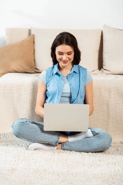 Freelancer sorrindo usando laptop no chão perto do sofá em casa — Fotografia de Stock