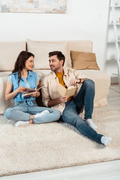 Mujer sonriente mirando al novio mientras lee libro sobre alfombra en casa - foto de stock