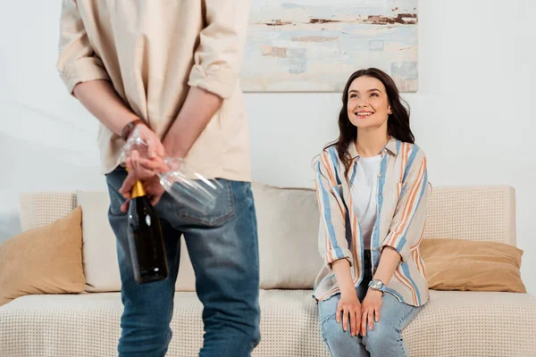 Enfoque selectivo de chica sonriente mirando novio escondiendo botella de champán y copas en casa - foto de stock