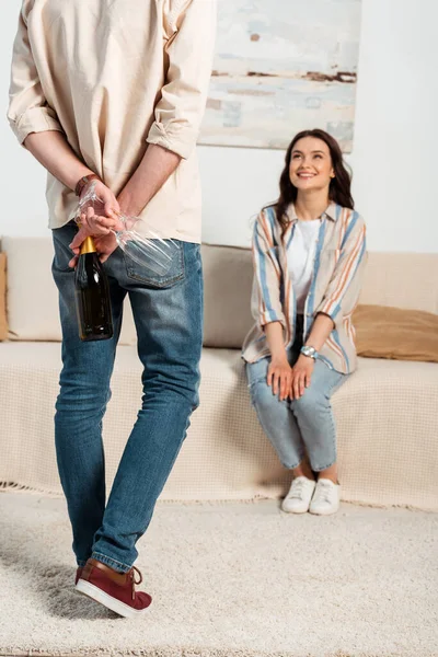 Выборочный фокус мужчины, прячущего бутылку шампанского и очки возле улыбающейся женщины на диване — стоковое фото