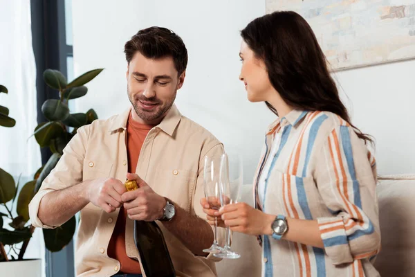 Hombre guapo abriendo botella de champán cerca de la mujer sosteniendo vasos en casa - foto de stock