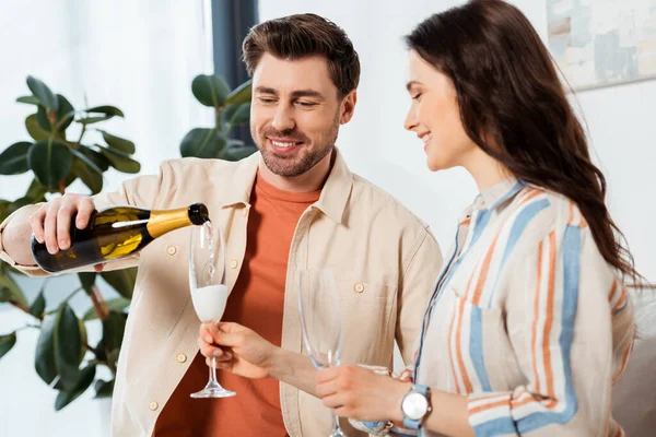 Выборочный фокус улыбающегося мужчины, наливающего шампанское в бокал рядом с красивой девушкой дома — стоковое фото