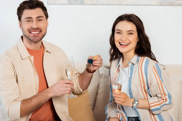 Casal alegre sorrindo para a câmera enquanto segurando copos de champanhe e chaves da nova casa no sofá — Fotografia de Stock