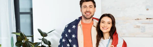 Récolte panoramique de couple souriant enveloppant dans le drapeau américain en regardant la caméra à la maison — Photo de stock