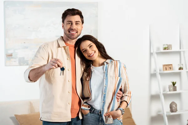 Jovem casal sorrindo para a câmera enquanto segurando as chaves da nova casa na sala de estar — Fotografia de Stock