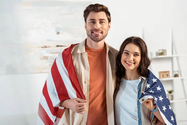 Выборочный фокус улыбающейся пары, смотрящей в камеру и заворачивающей американский флаг — стоковое фото