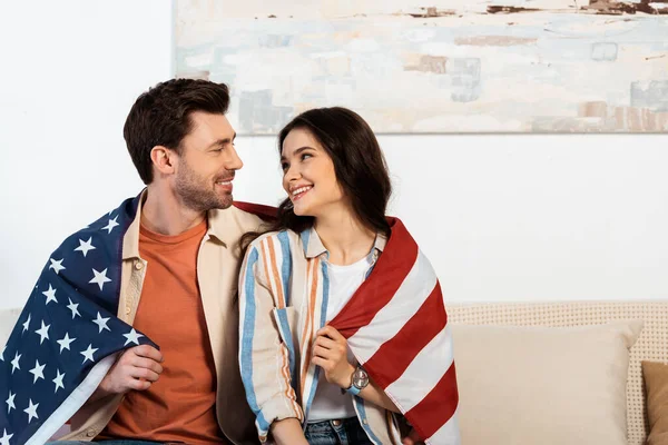 Молода пара, загорнута в американський прапор посміхаючись один одному вдома — стокове фото