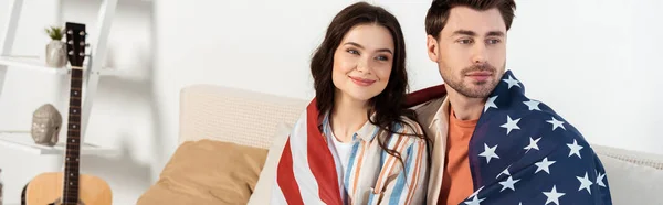 Panoramic shot of smiling woman wrapped in american flag sitting near boyfriend on couch — Stock Photo