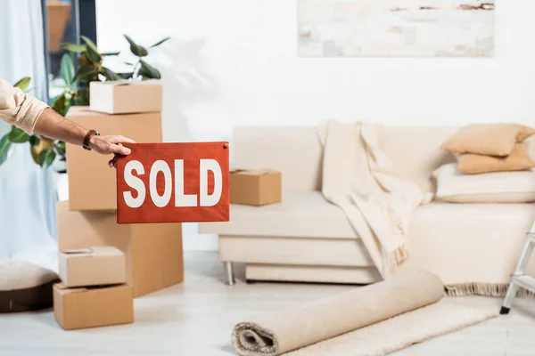 Cropped view of man holding nameplate with sold lettering near cardboard boxes in living room at background — Stock Photo