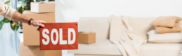 Panoramic crop of man holding nameplate with sold lettering with cardboard boxes in living room at background — Stock Photo