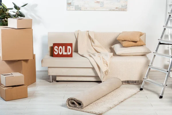 Nameplate with sold lettering on couch near ladder and cardboard boxes in living room — Stock Photo