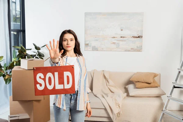 Mujer joven sosteniendo placa con letras vendidas cerca de cajas de cartón en la sala de estar - foto de stock