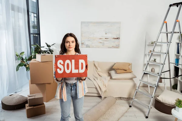 Attrayant fille tenant plaque signalétique avec lettrage vendu près de boîtes en carton et échelle dans le salon — Photo de stock