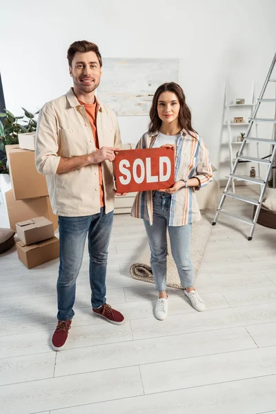 Smiling couple holding nameplate with sold lettering with cardboard boxes and ladder at background — Stock Photo