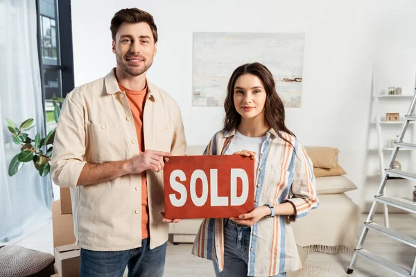 Smiling man holding nameplate with sold lettering near girlfriend in living room — Stock Photo