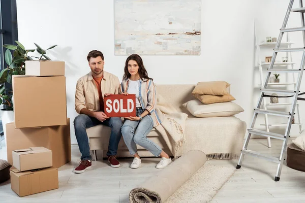 Couple bouleversé tenant plaque signalétique avec lettrage vendu près de boîtes en carton et échelle à la maison — Photo de stock