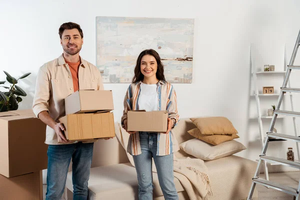 Young couple smiling at camera and holding cardboard boxes in living room during relocation — Stock Photo