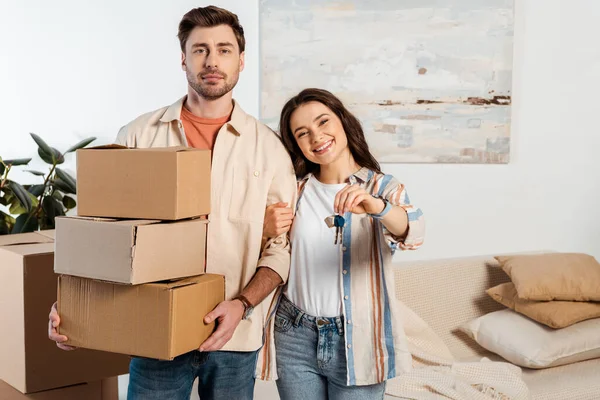 Femme souriante tenant les clés de la nouvelle maison à côté du petit ami tenant des boîtes en carton à la maison — Photo de stock