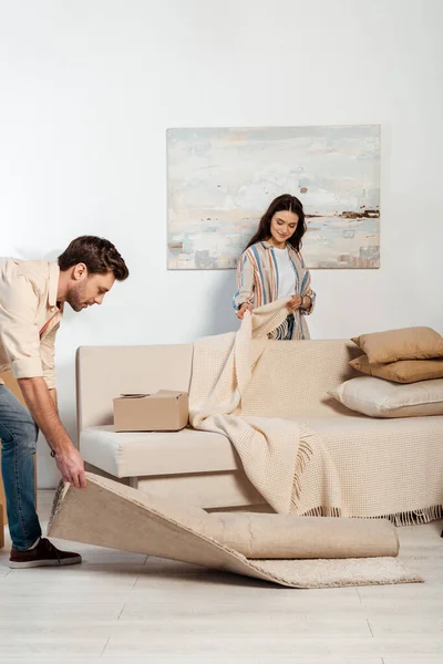 Selective focus of woman holding plaid near couch while boyfriend turning off carpet in living room — Stock Photo