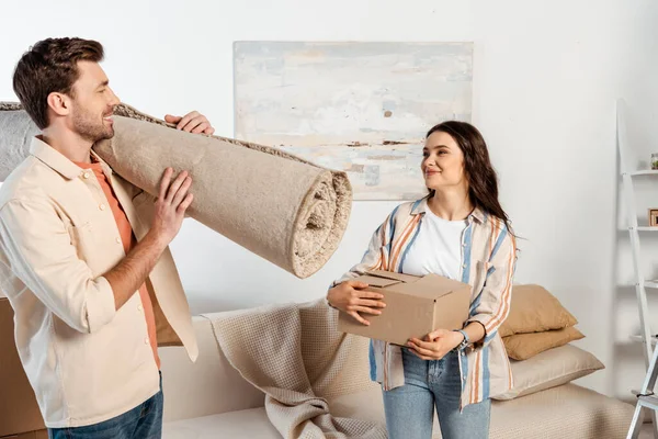 Young man holding carpet and smiling at girlfriend with cardboard box in living room — Stock Photo