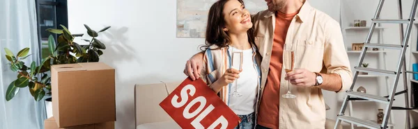 Panoramic shot of smiling woman holding glass of champagne near boyfriend with sold lettering on nameplate in living room — Stock Photo