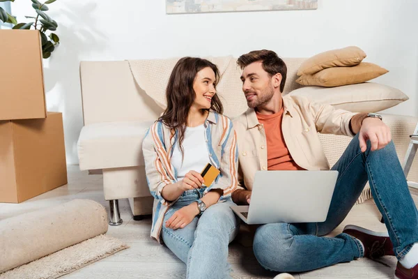 Couple souriant utilisant ordinateur portable et carte de crédit sur le sol pendant le déménagement — Stock Photo