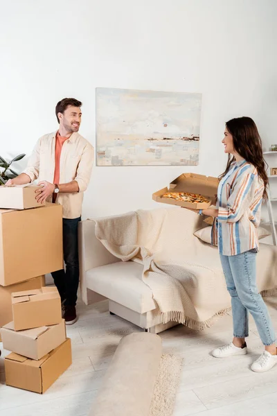 Smiling man staking cardboard boxes while girlfriend holding pizza box at home — Stock Photo