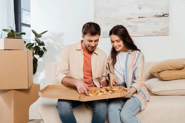 Sorrindo casal segurando caixa de pizza no sofá na casa nova — Fotografia de Stock