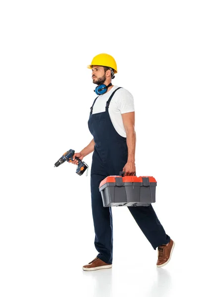 Handsome workman in overalls and hardhat holding toolbox and electric screwdriver while walking on white background — Stock Photo