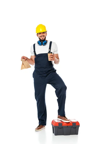 Smiling repairman in uniform holding paper bag and disposable cup near toolbox on white background — Stock Photo