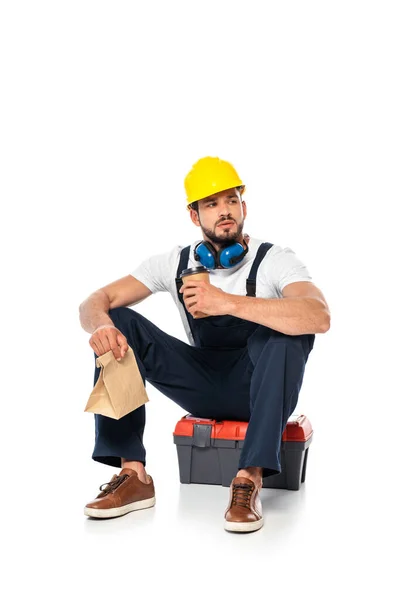 Handsome workman holding coffee to go and paper bag while sitting on toolbox on white background — Stock Photo