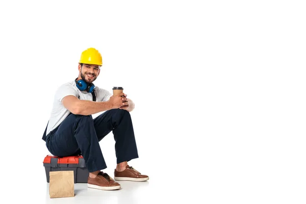 Reparador sonriente en uniforme sosteniendo café para llevar mientras está sentado en la caja de herramientas cerca de la bolsa de papel sobre fondo blanco — Stock Photo