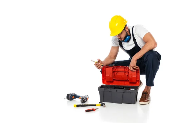 Trabalhador em hardhat e defensores de orelha segurando chave de fenda perto de ferramentas e caixa de ferramentas no fundo branco — Fotografia de Stock